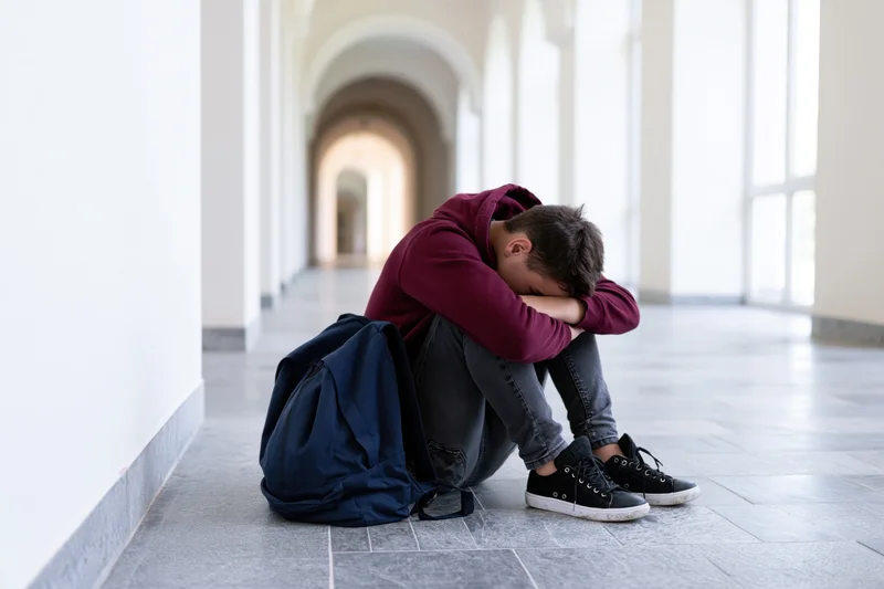 Teenage boy sitting on floor in school hallway with head in hands, backpack beside him