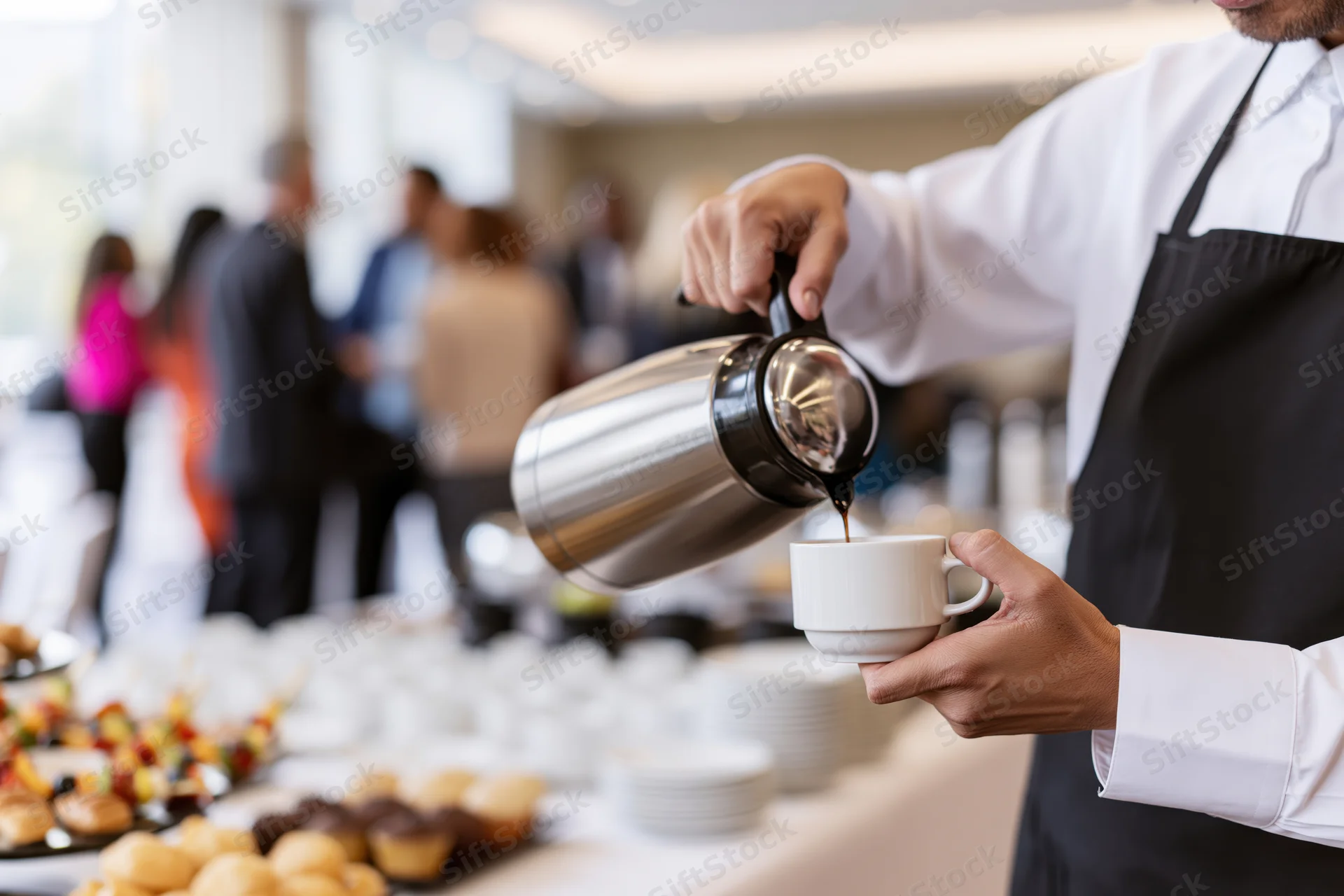 Waiter pouring coffee from a thermos into a cup at a buffet table with pastries and guests in the background