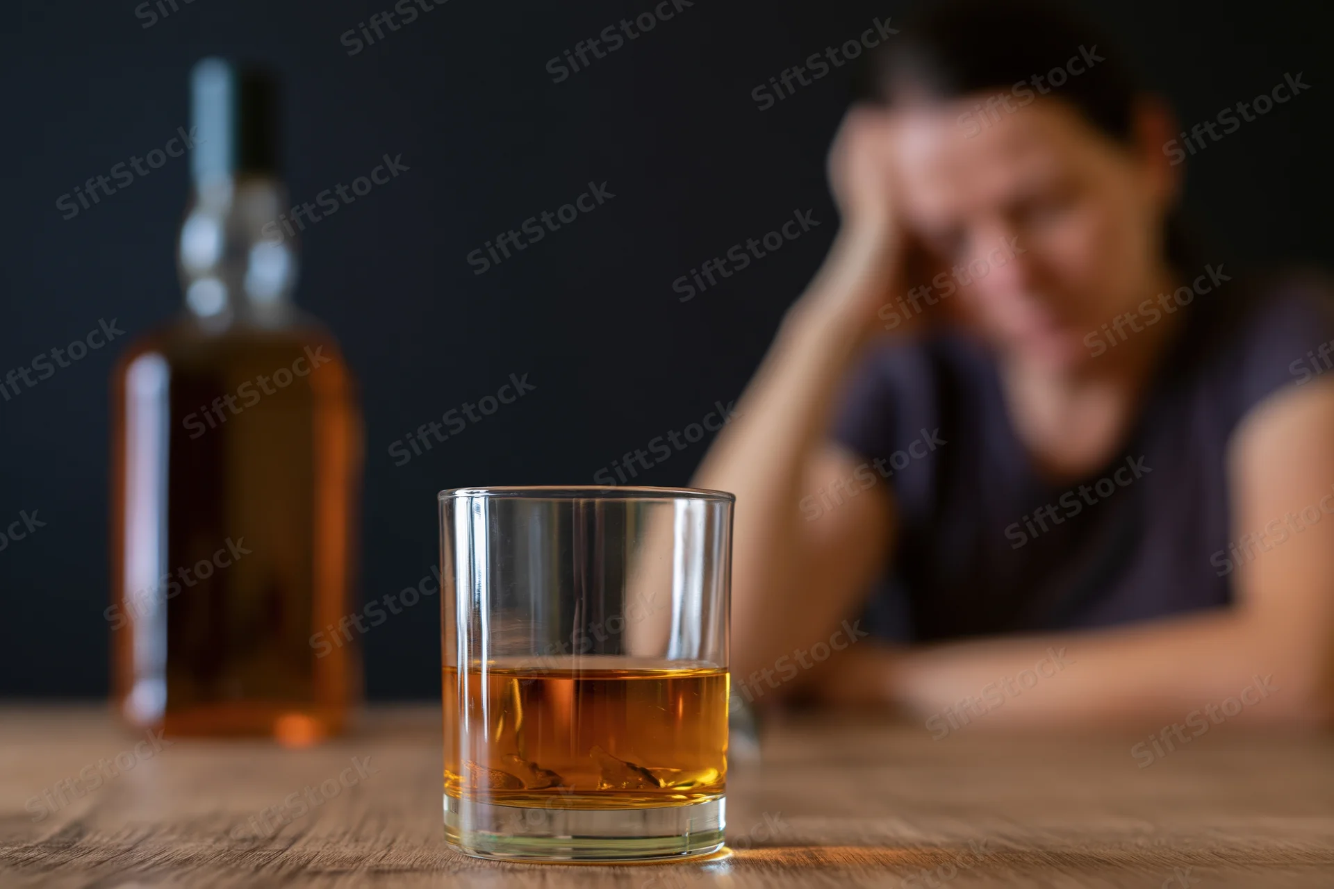 Whiskey glass and bottle in focus on wooden table with distressed woman in background