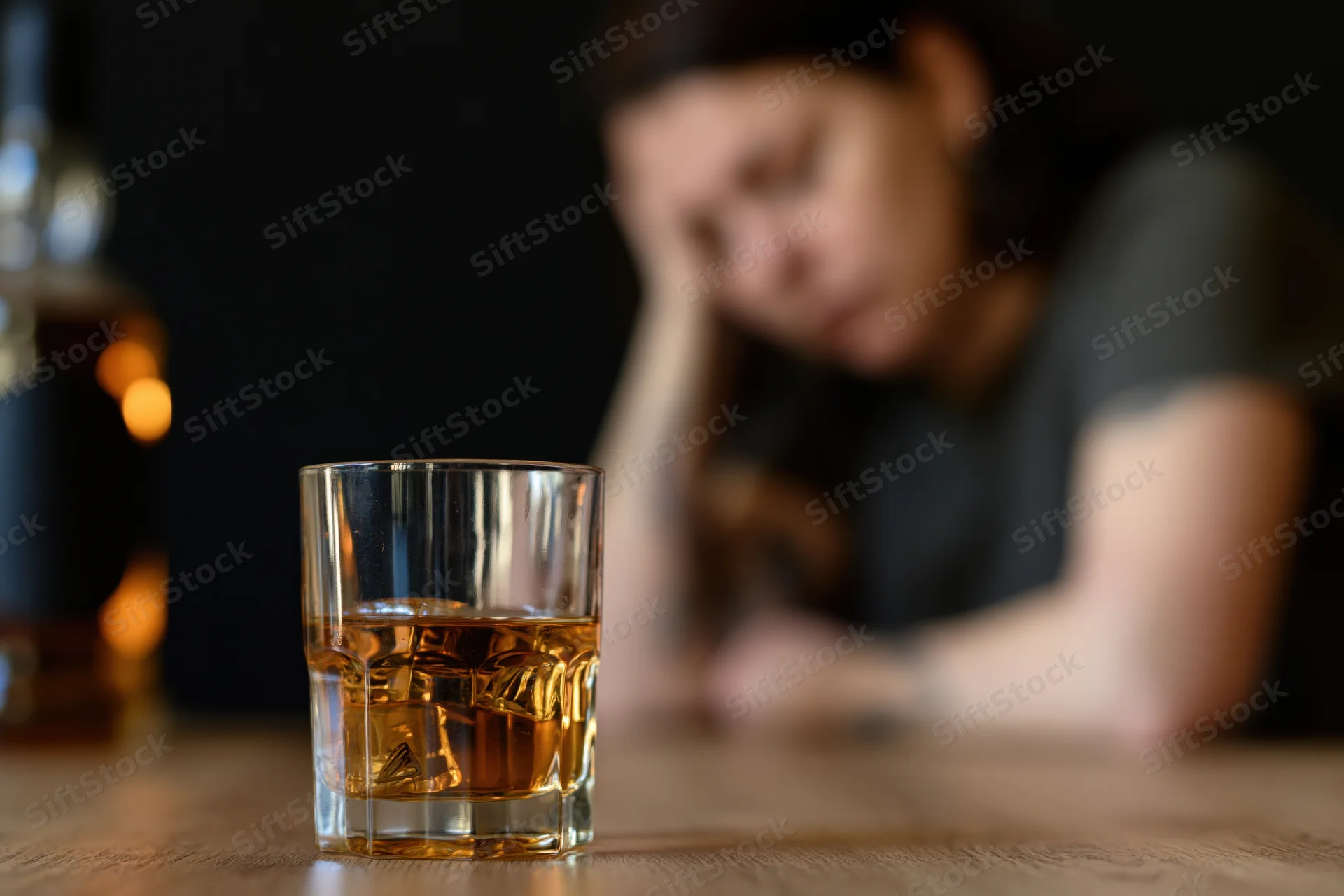 Whiskey glass with ice on wooden table, woman in background resting head on hand