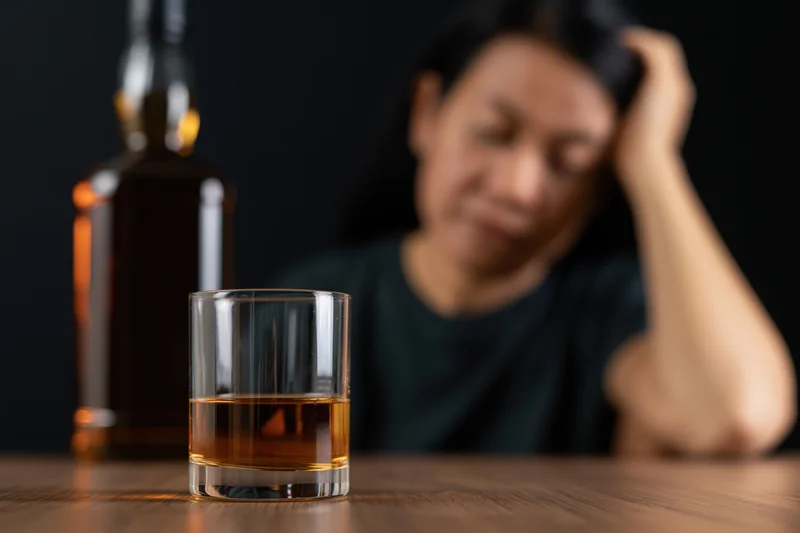 Whiskey glass and bottle in focus on wooden table with blurred woman in background holding head