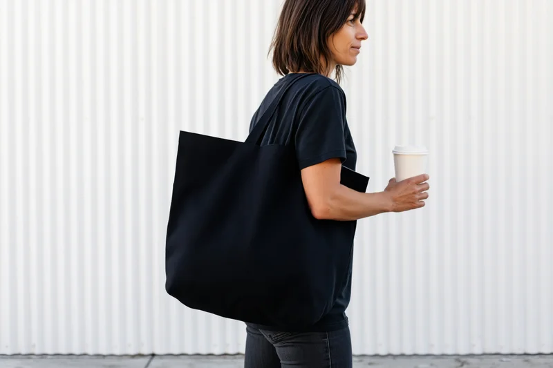 Woman carrying black canvas tote bag over shoulder while walking outdoors holding coffee cup