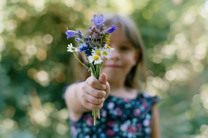 Young girl holding small wildflower bouquet toward camera in sunlit garden