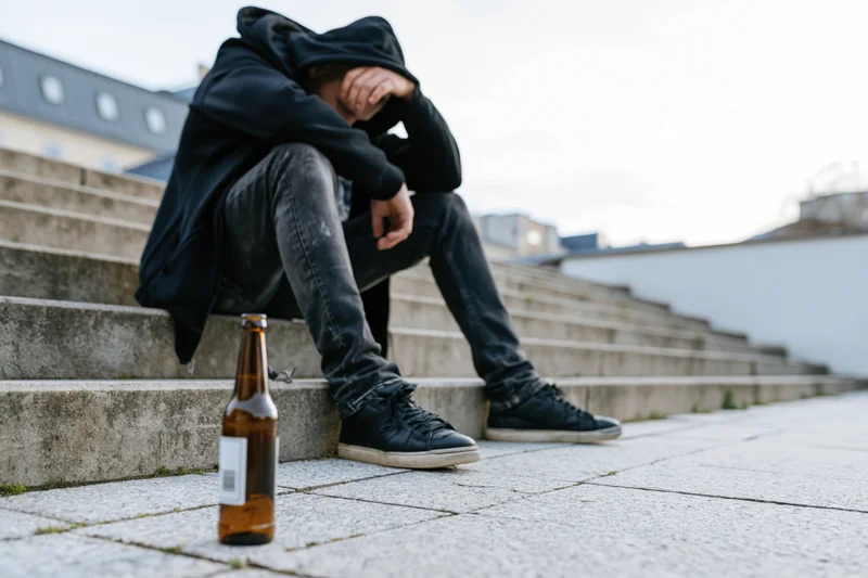 Young man in black hoodie sitting slumped on concrete steps with beer bottle nearby