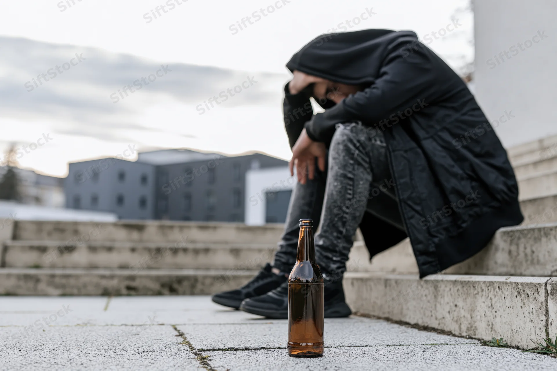 Young man in black hoodie slumped on concrete steps with beer bottle nearby