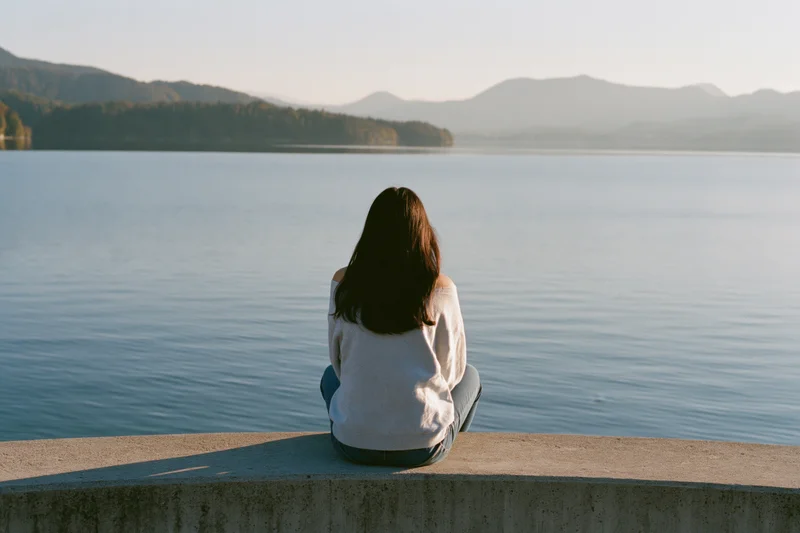 Young woman sitting alone on concrete ledge facing calm lake with mountains in distance