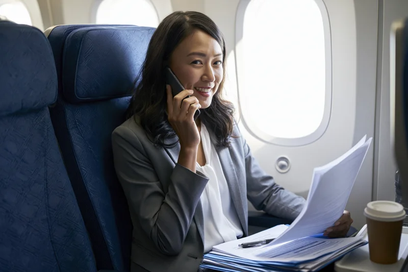 Asian businesswoman talking on phone while reviewing documents in airplane cabin