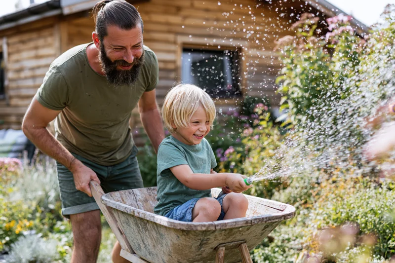 Bearded father pushing wheelbarrow with smiling blonde son holding sprinkler in garden