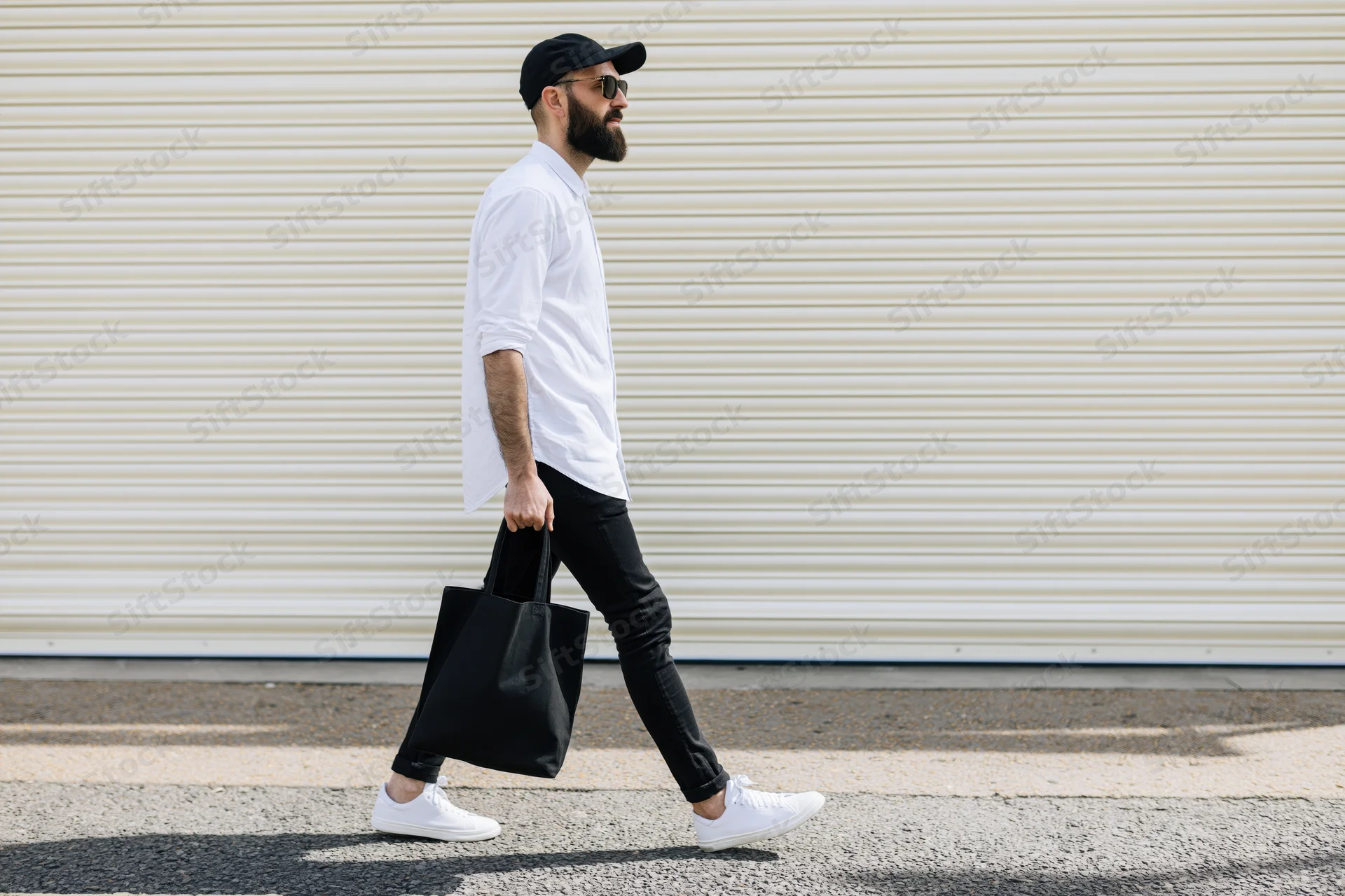 Bearded man in white shirt and black cap walking on sidewalk carrying tote bag