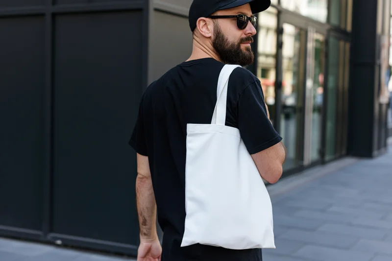 Bearded young man in black cap and sunglasses carrying white tote bag on city sidewalk