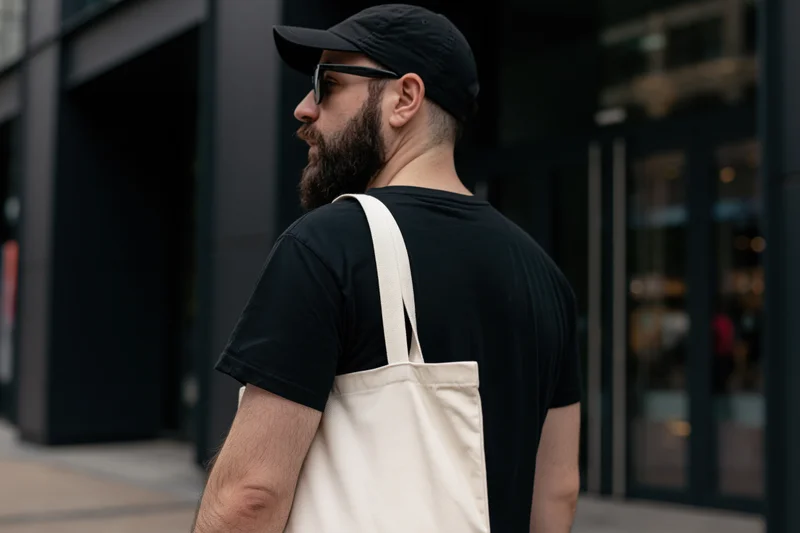 Bearded young man in black cap and sunglasses carrying tote bag on city sidewalk