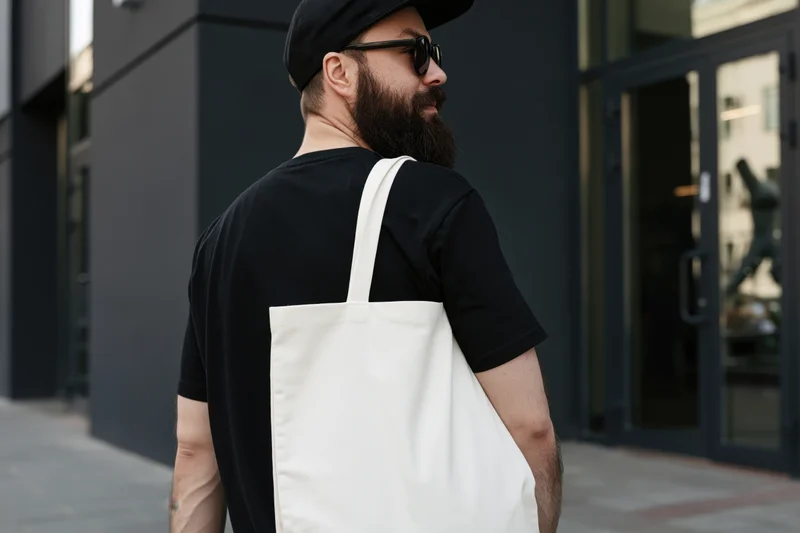 Bearded young man in black cap and sunglasses standing on city sidewalk with white tote bag over shoulder
