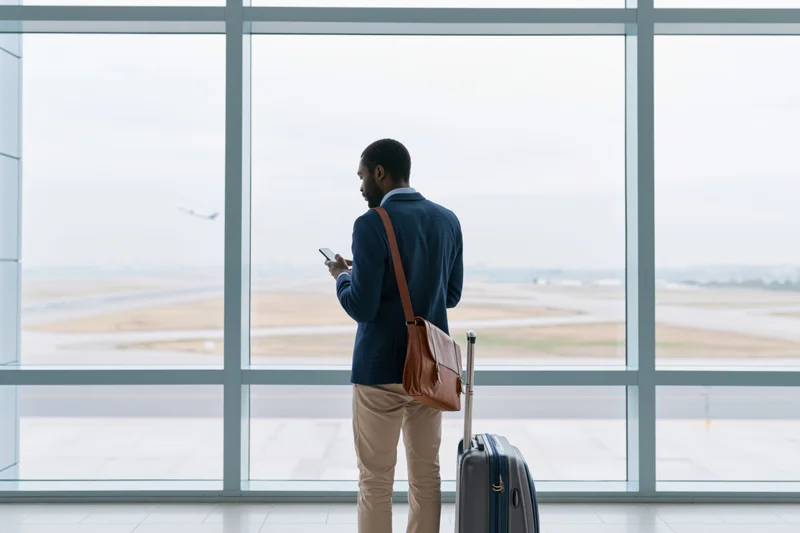 Businessman standing at airport terminal window holding smartphone with suitcase beside him