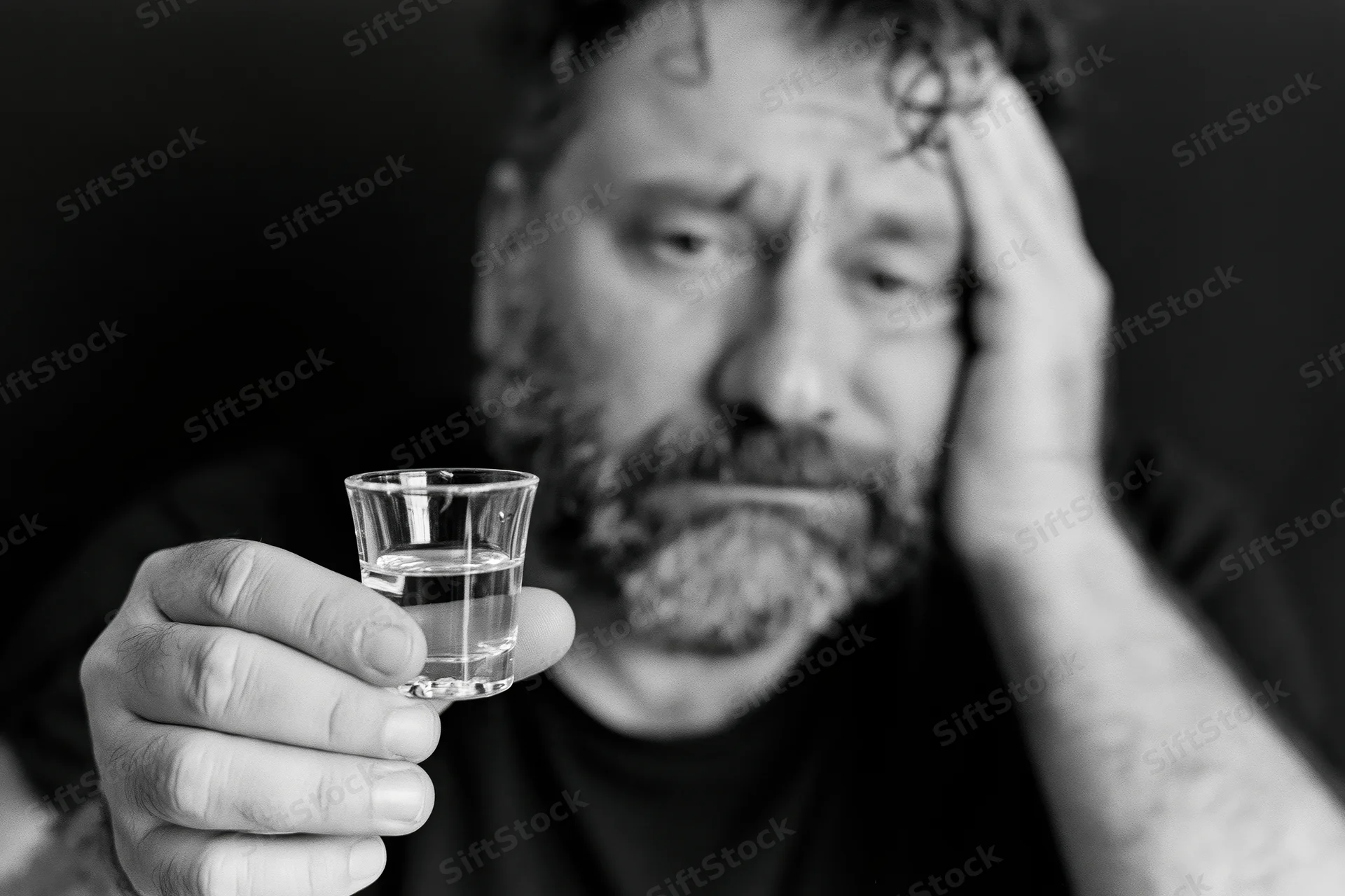 Distressed bearded man holding a shot glass of clear liquor, hand on head, dark background