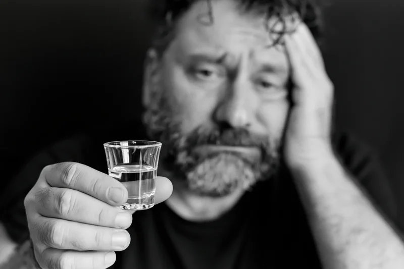 Distressed bearded man holding a shot glass of clear liquor, hand on head, dark background