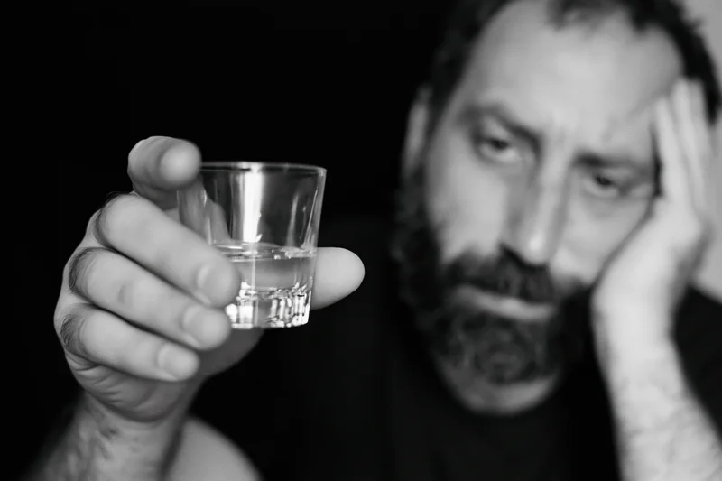 Distressed bearded man holding a shot glass of clear liquid, hand on head, in shadowed closeup