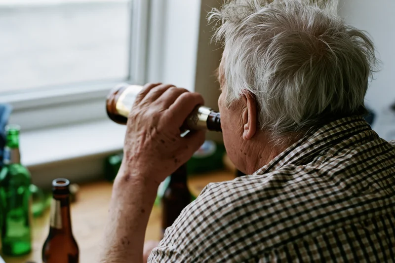 Elderly man drinking from a brown beer bottle at a table by a window