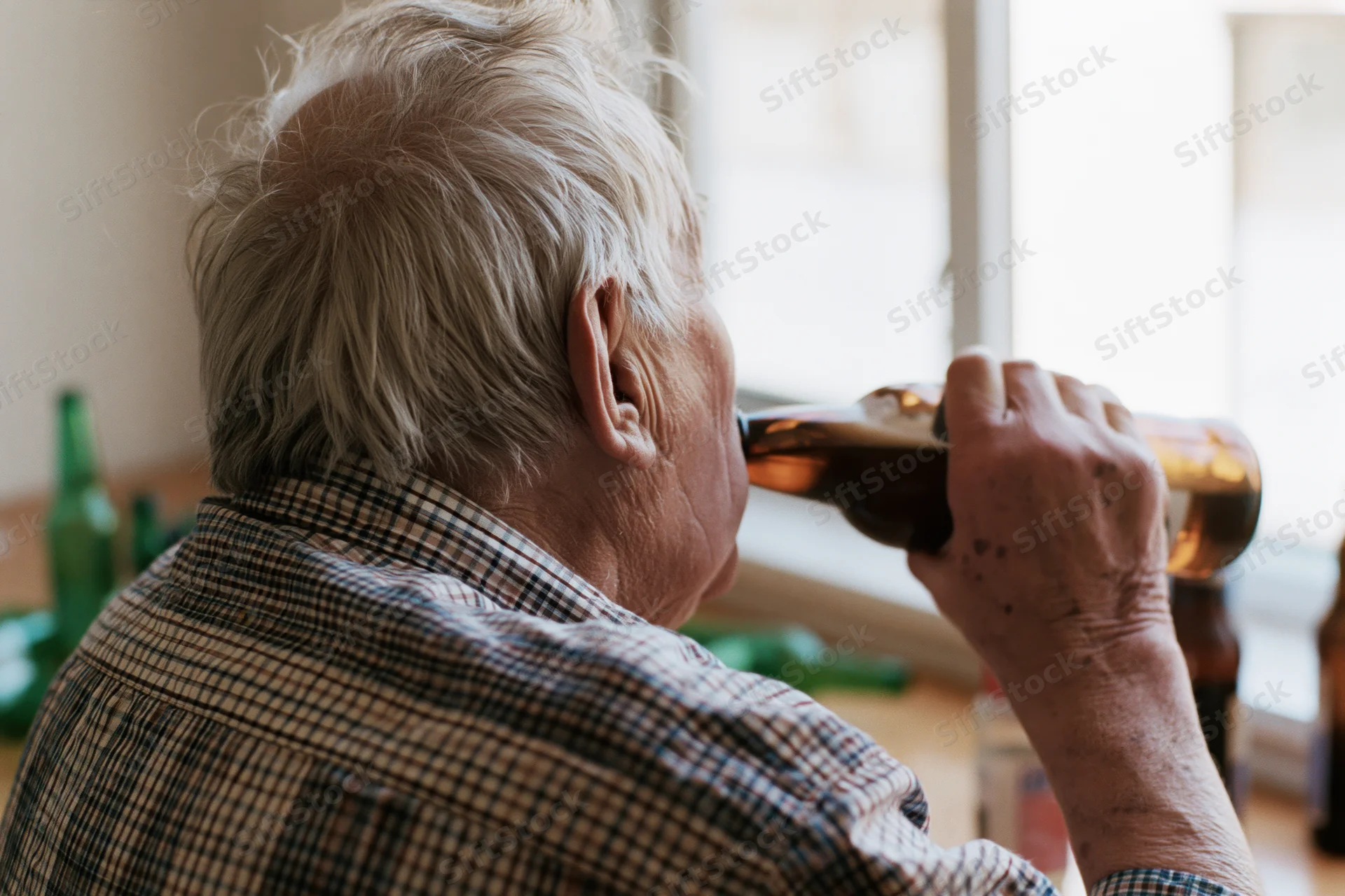 Elderly man drinking from a brown beer bottle indoors near a window