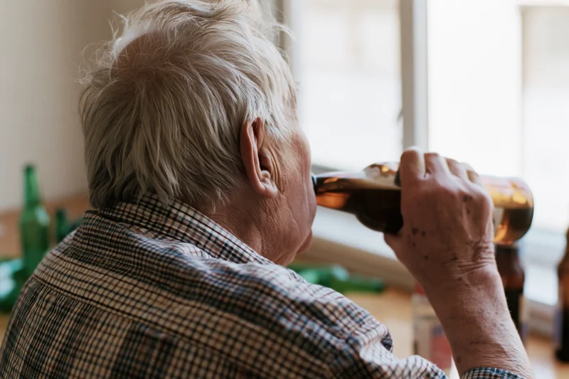 Elderly man drinking from a brown beer bottle indoors near a window