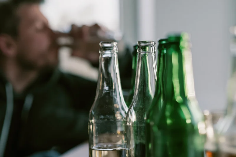 Empty glass beer bottles in focus on a table with a blurred man drinking in the background