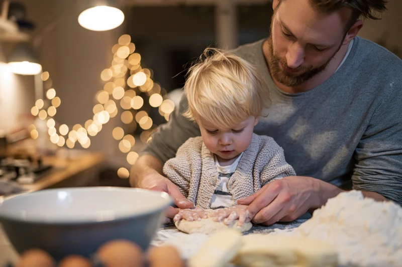 Father and young blonde toddler baking dough together in kitchen with holiday lights in background