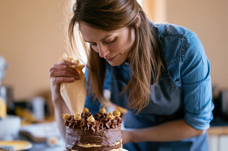 Woman decorating a layered chocolate caramel cake with a piping bag in a kitchen