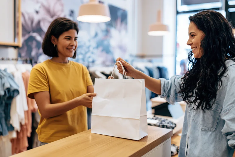 Woman shop assistant handing white paper bag to smiling customer at clothing store counter