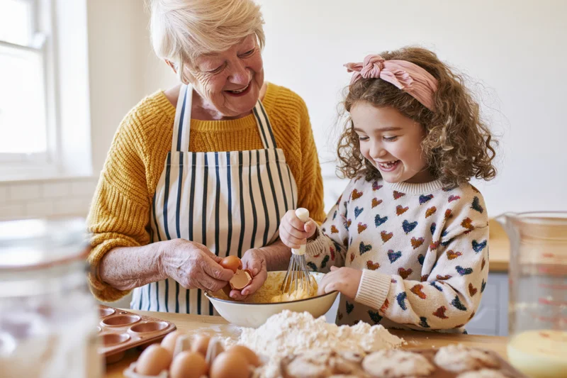 Grandmother and granddaughter baking together in a kitchen, smiling over a mixing bowl with flour and eggs on the counter