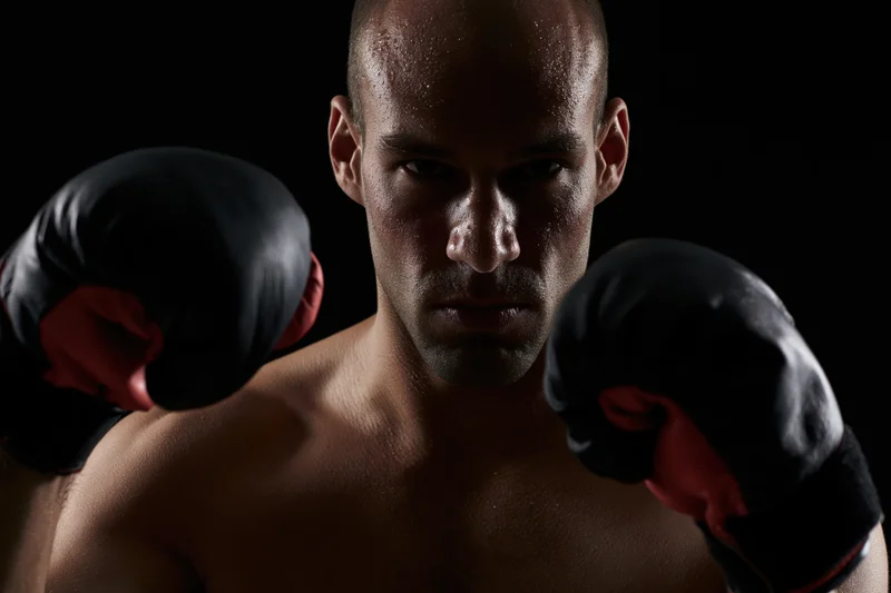 Male boxer in fighting stance with fists raised, shirtless and sweating, wearing black and red gloves against dark background