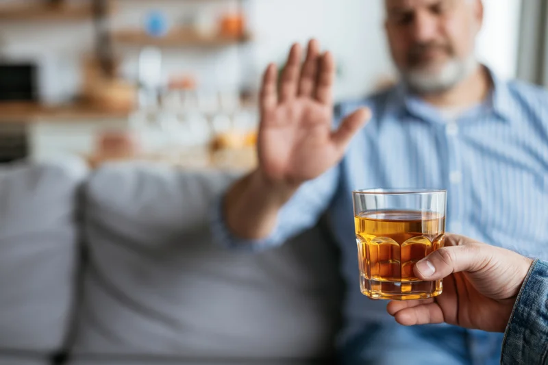 Man raising palm in refusal gesture toward a glass of whiskey held by another person in a living room