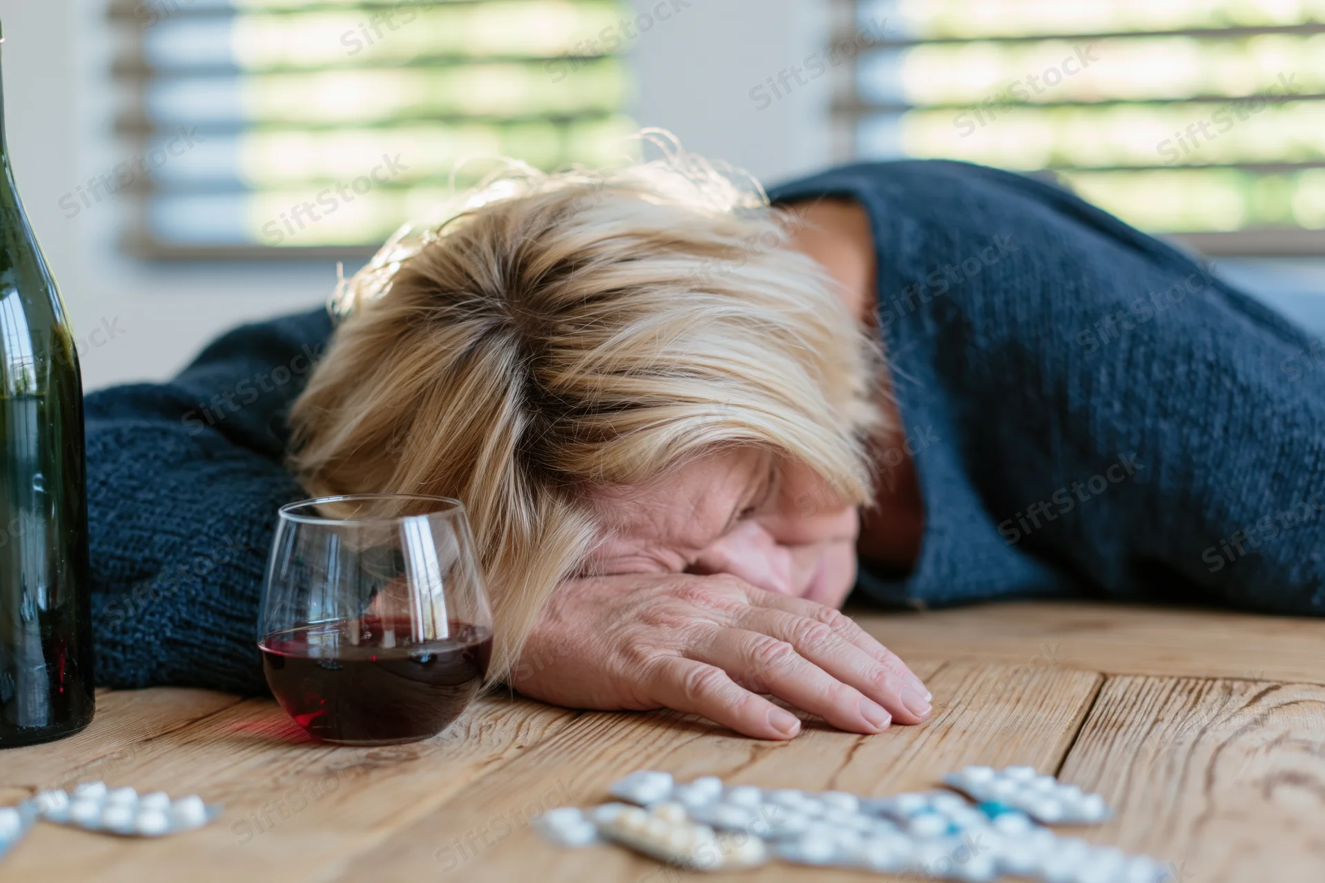 Middle-aged blonde woman slumped face-down on wooden table with wine glass and scattered pills