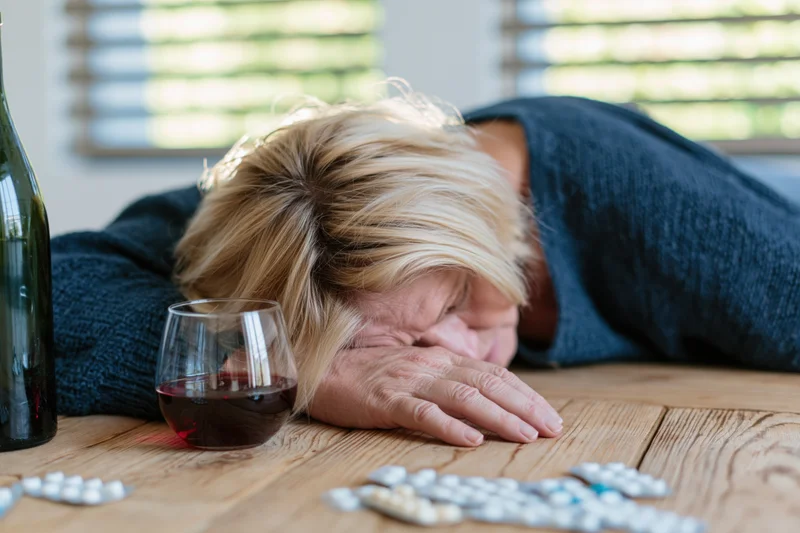 Middle-aged blonde woman slumped face-down on wooden table with wine glass and scattered pills