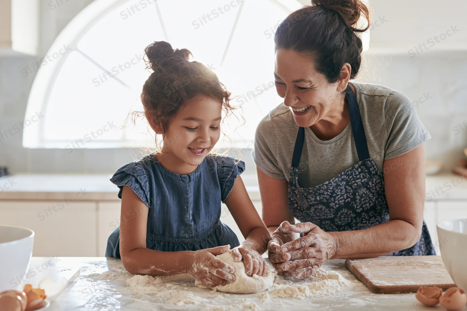 Mother and daughter laughing while kneading dough together in a bright kitchen