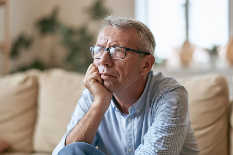Senior man with glasses sitting thoughtfully on a beige sofa in a living room