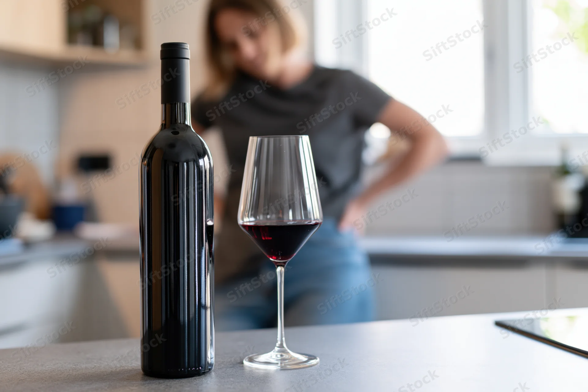 Bottle of red wine and a half-full glass on a kitchen countertop with a woman in the background