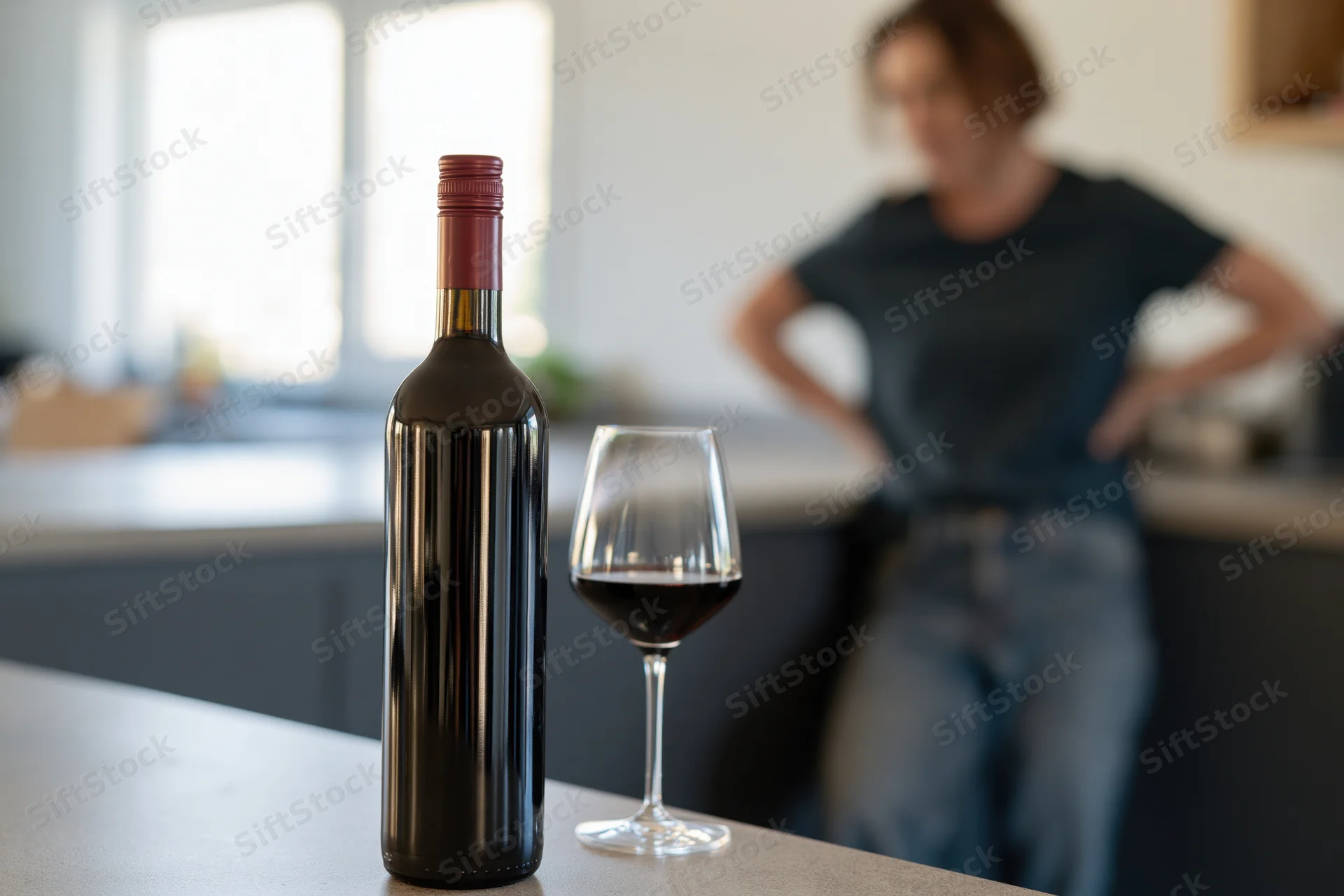 Bottle of red wine and a half-full glass on a kitchen countertop with a woman in the background