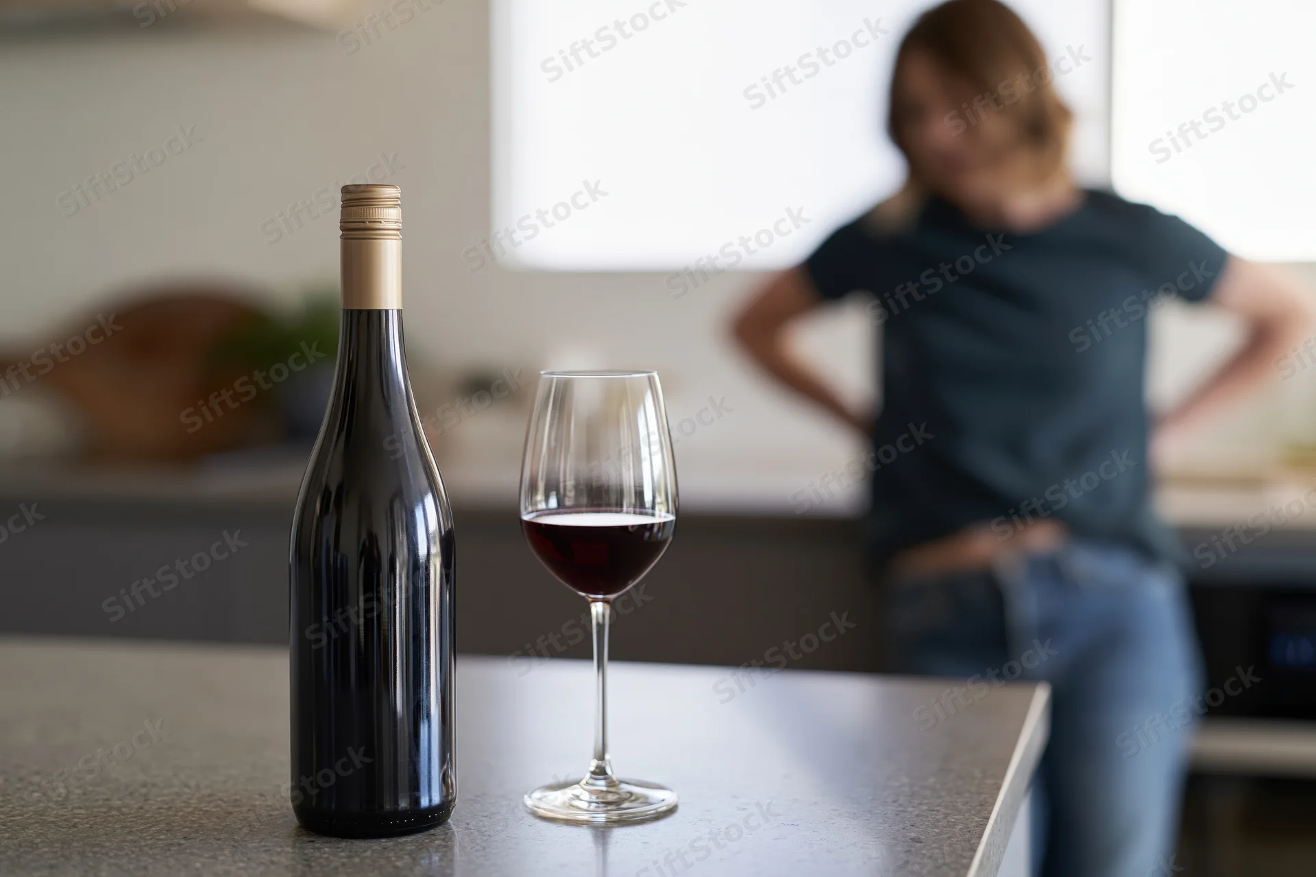 Bottle of red wine and a half-full glass on a kitchen countertop with a woman standing blurred in the background