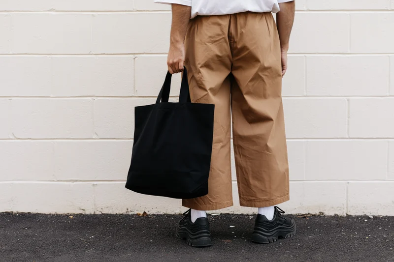 Man holding black tote bag against white brick wall, back view, casual streetwear outfit