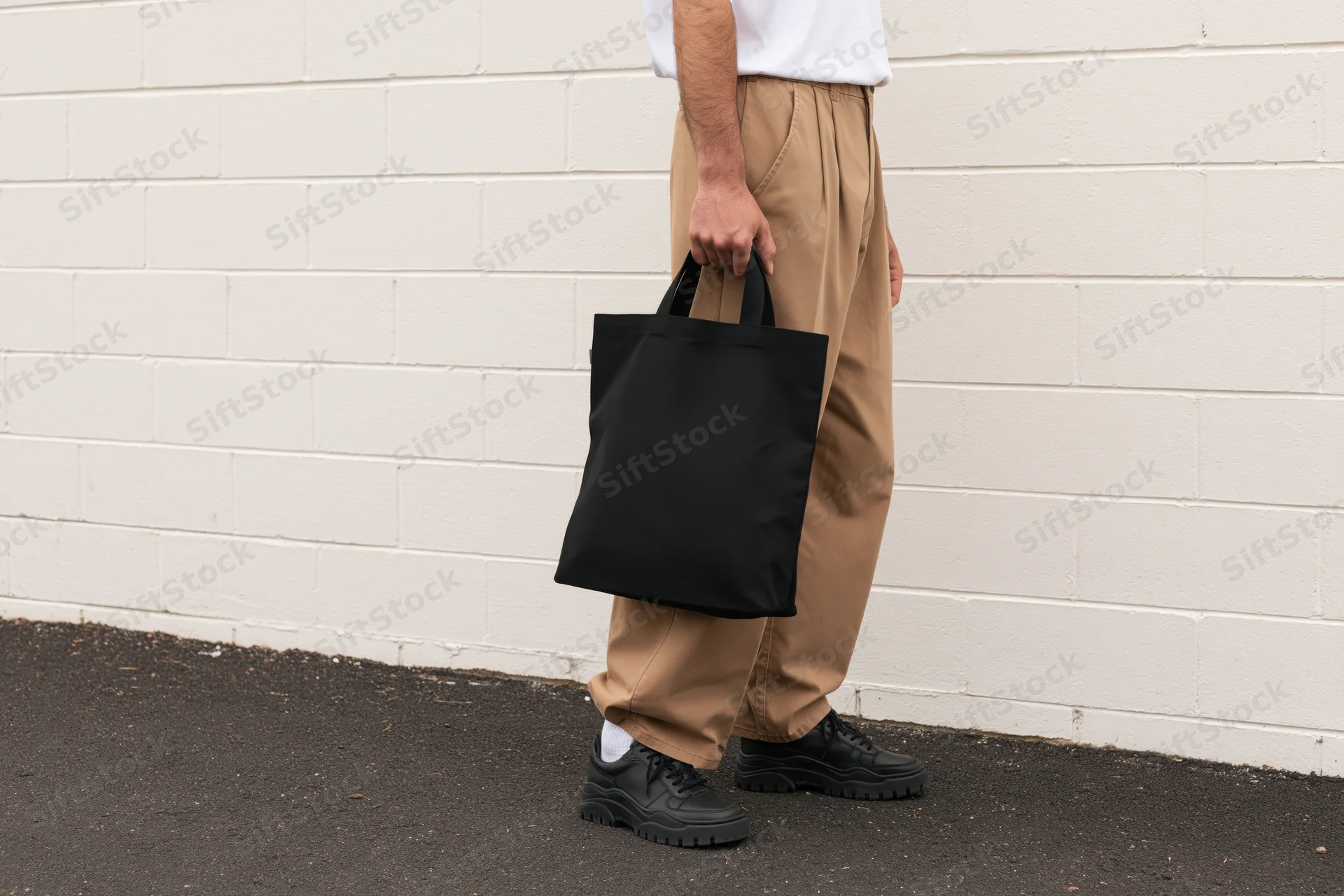 Man holding black tote bag against white brick wall outdoors