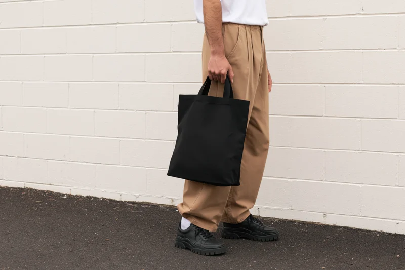 Man holding black tote bag against white brick wall outdoors