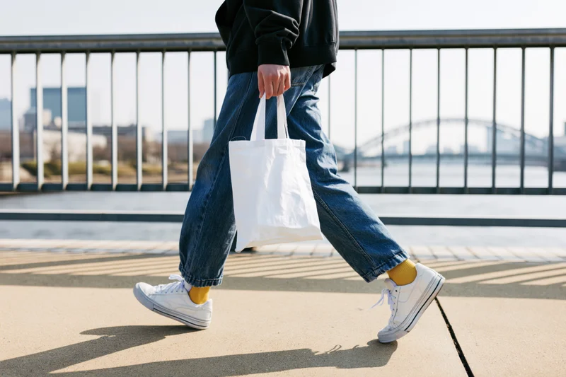 Person walking across pedestrian bridge holding white tote bag, wearing jeans and sneakers, urban setting