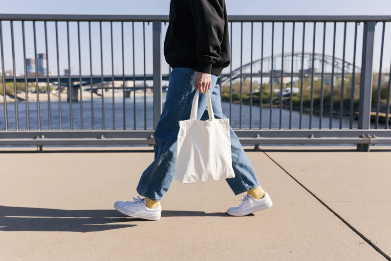 Woman walking across pedestrian bridge holding white tote bag, wearing jeans and sneakers, urban riverside setting