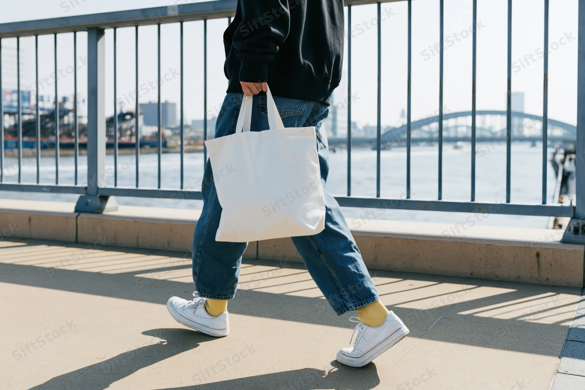 Person walking across pedestrian bridge holding white tote bag by waterfront