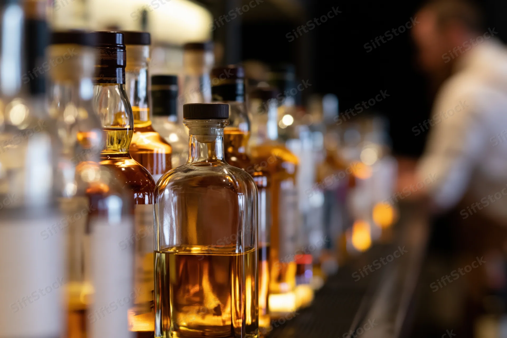 Row of whiskey bottles lined up on a backlit bar shelf with a bartender in the background