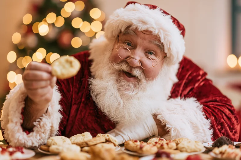 Santa holding a cookie in front of a decorated christmas tree at a festive table