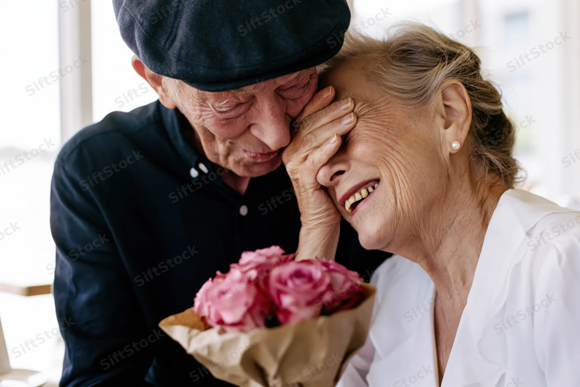 Senior man in flat cap covering his smiling wife’s eyes with a bouquet of pink roses indoors