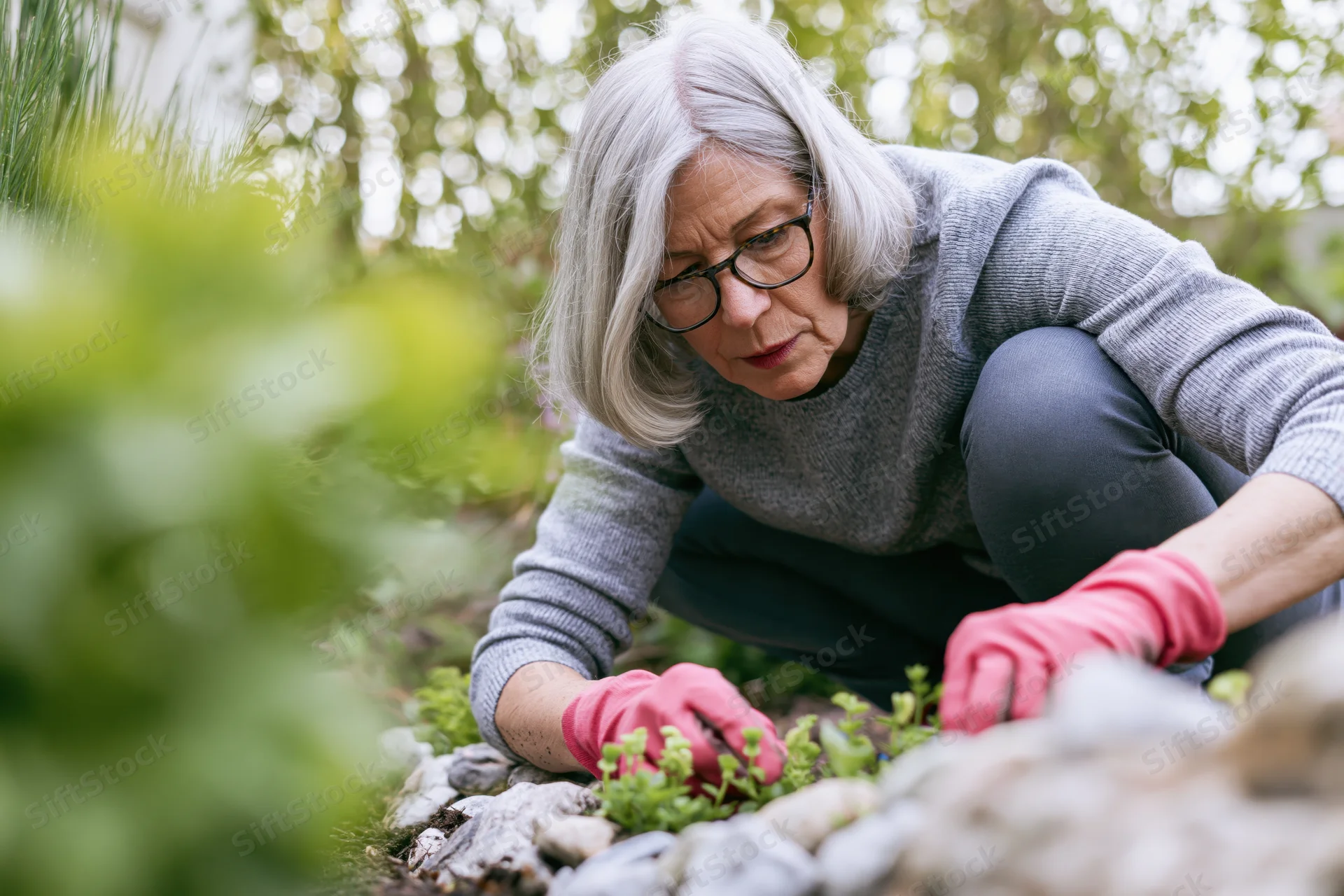 Senior woman crouching to tend small plants in a garden bed surrounded by rocks and greenery