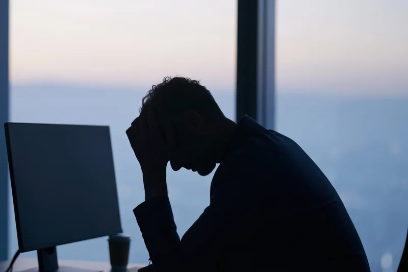 Businessman in silhouette at desk with head in hands near computer monitor by window at twilight
