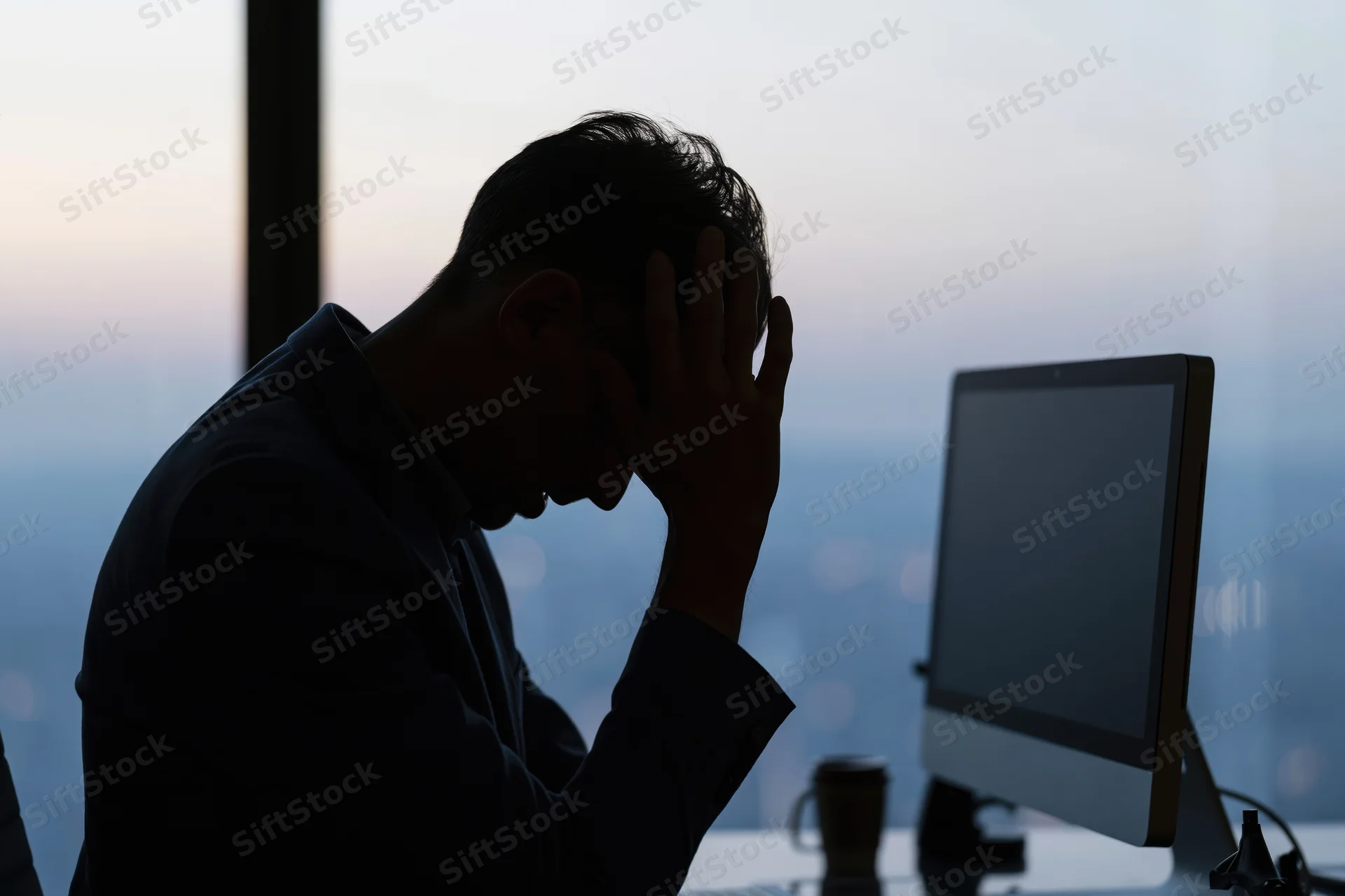 Businessman in silhouette holding head at desk in front of computer during twilight