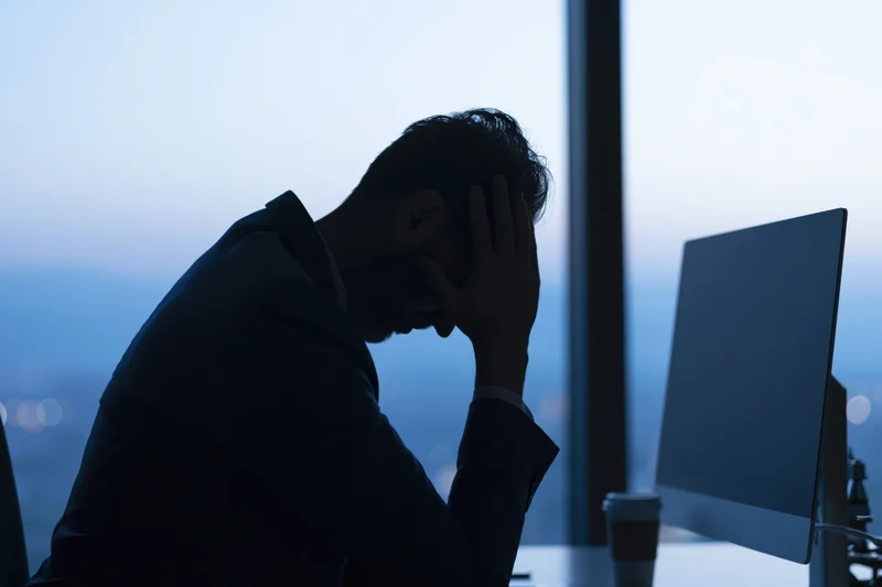Businessman in silhouette at desk with head in hands, office at twilight